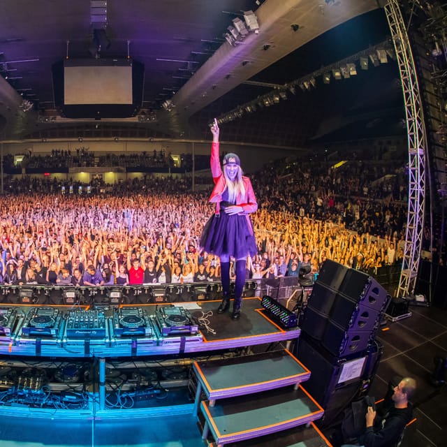 CANDICE standing on the DJ booth with one arm raised before a packed indoor arena