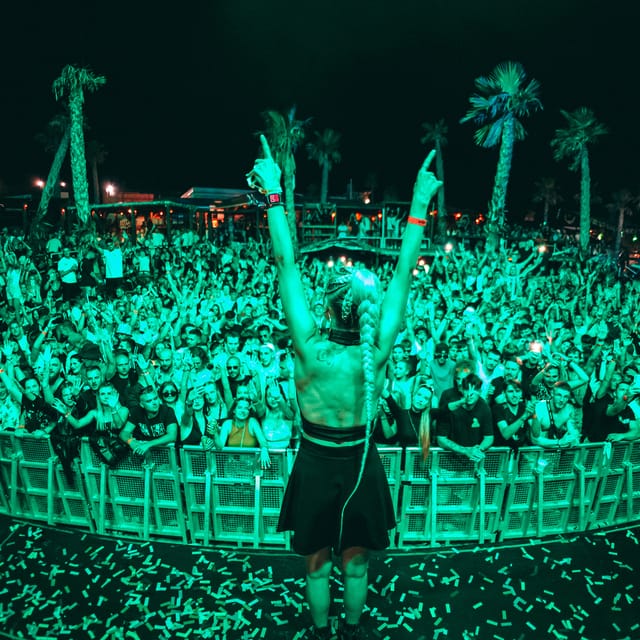 CANDICE facing a packed outdoor crowd with both arms raised under green stage lighting
