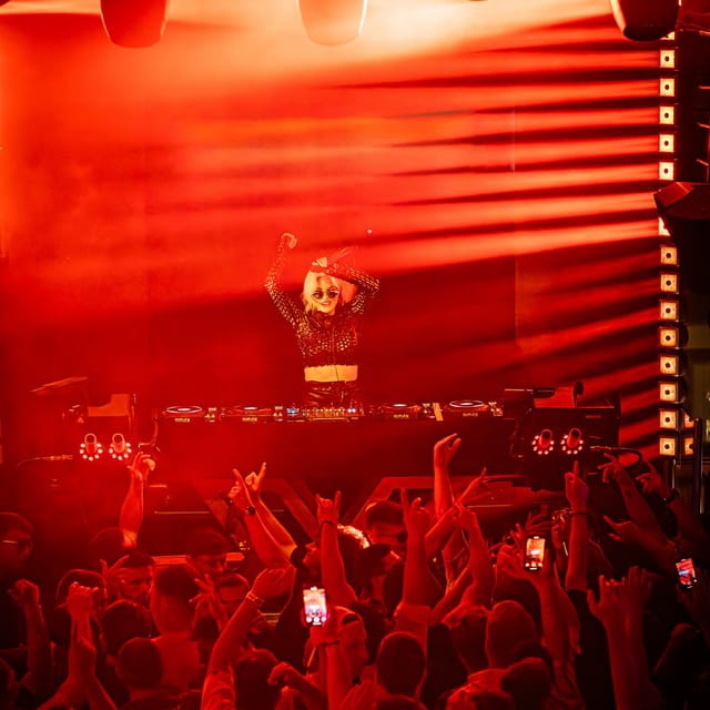 CANDICE raising her hands behind the DJ booth under intense red club lighting
