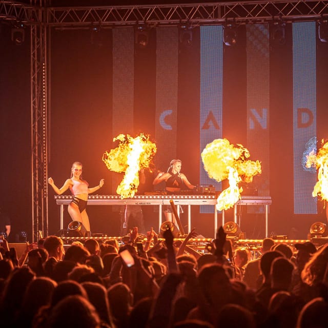 CANDICE performing between two dancers as fire effects burst across an indoor stage