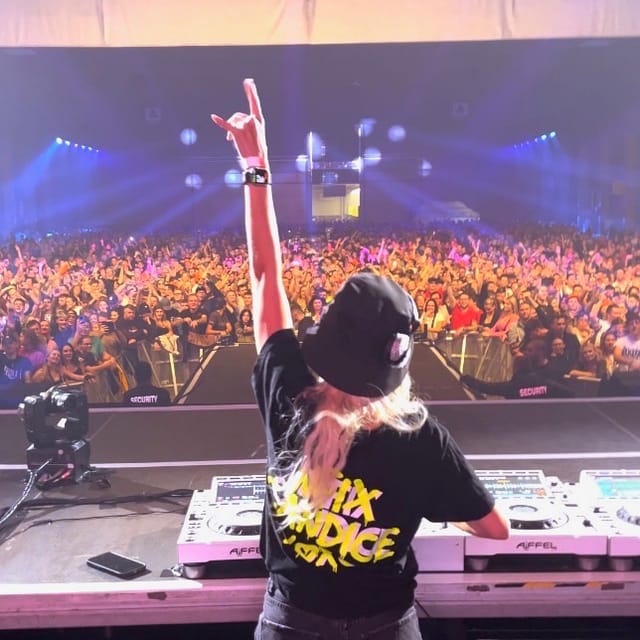 CANDICE raising one arm behind the DJ decks toward a packed indoor crowd