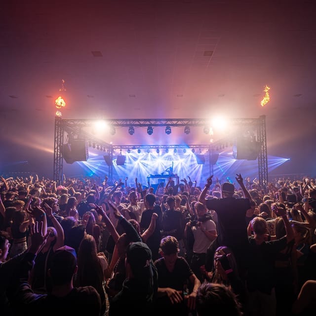 A packed indoor audience facing CANDICE beneath blue stage lights and flame effects