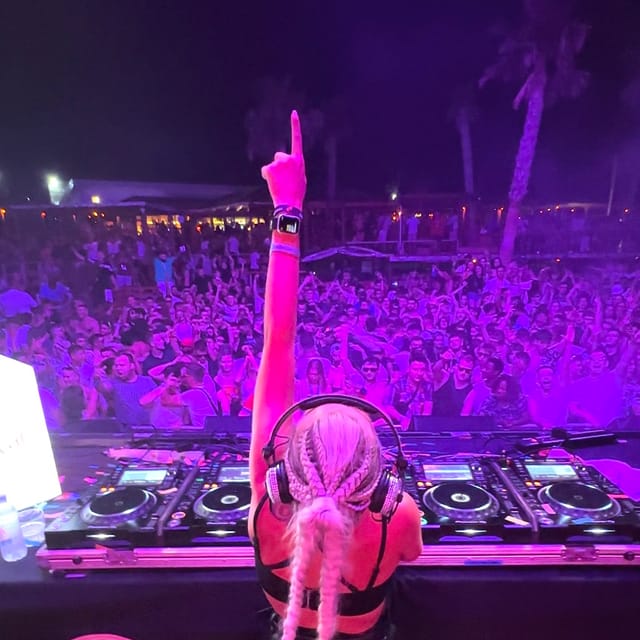 CANDICE pointing upward behind the DJ decks toward a purple-lit outdoor festival crowd